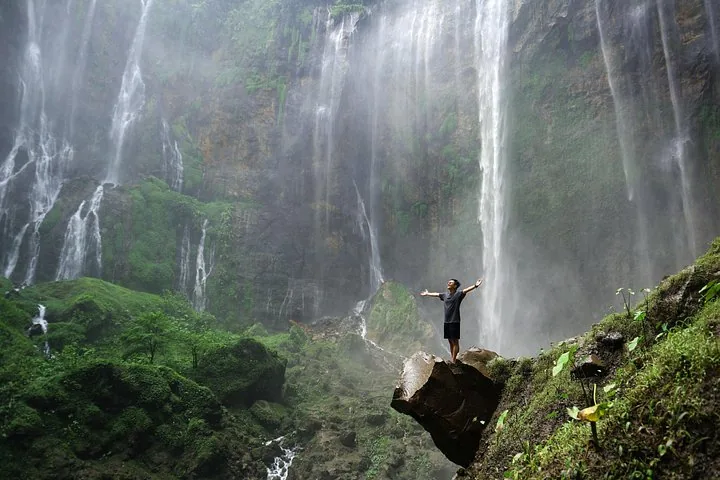 man standing on a rock with arms wide at the base of Tumpak Sewu Waterfall
